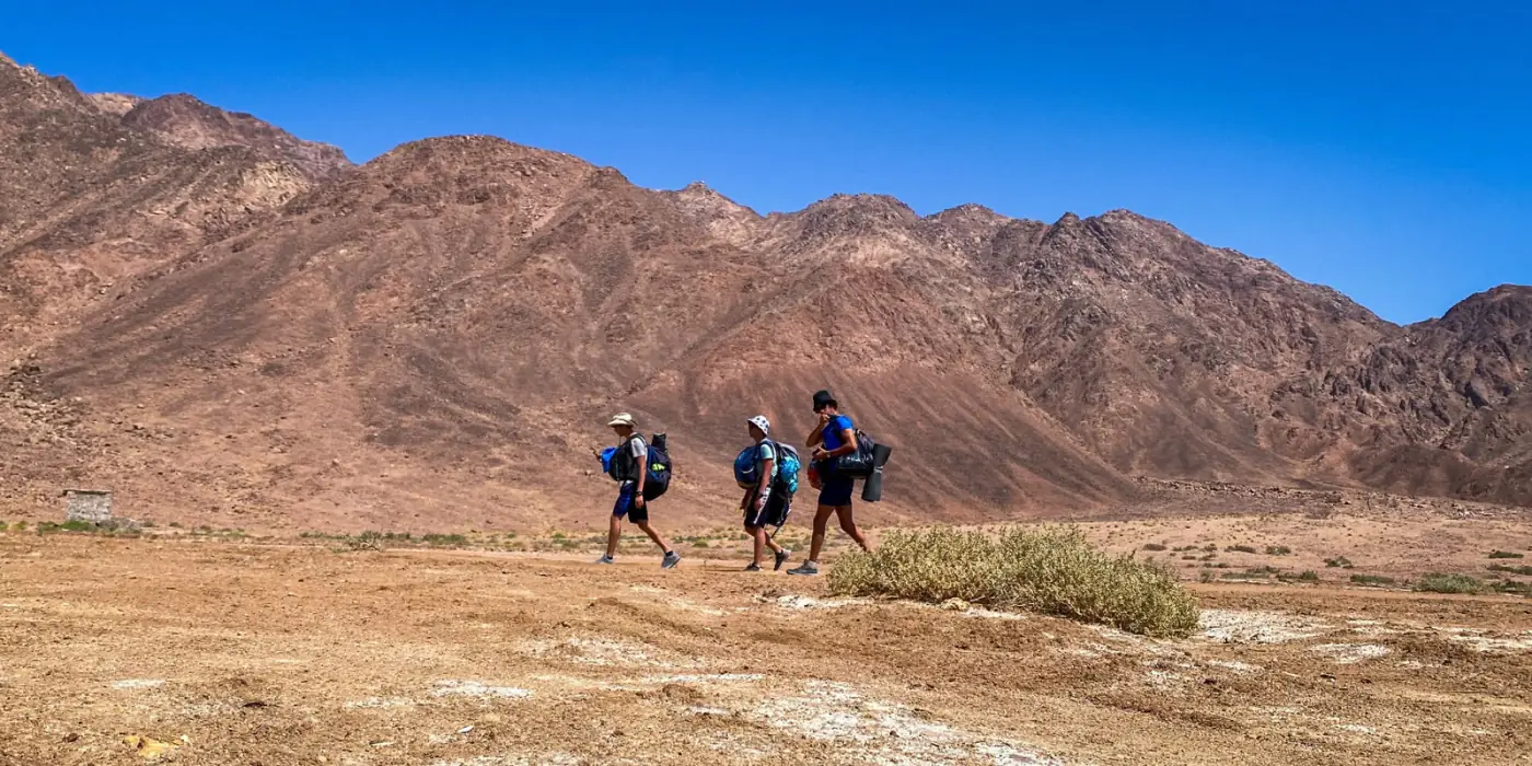Three hikers crossing a desert valley below a mountain ridge.
